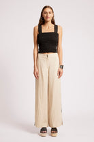 A woman modeling a black smocked sleeveless crop top and beige wide-leg trousers with side buttons, paired with black platform sandals, against a light gray studio background.