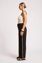 A woman shown in profile wearing black wide-leg pants with a light side stripe, a cream ribbed tank top, and black sandals against a plain light gray studio background.