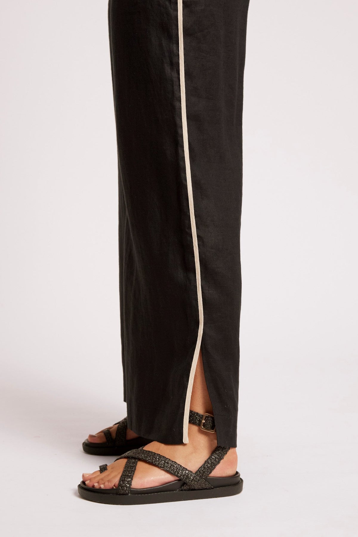 Black wide-leg trousers with a beige side stripe and a front slit, shown on a model wearing black strappy sandals against a light gray studio background.