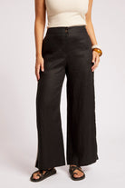 A model wearing black wide-leg linen pants with a button fly, styled with a cream sleeveless top and black sandals against a light gray studio background.