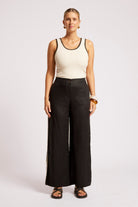 A woman stands against a light gray studio background wearing a cream sleeveless top with black trim and black wide-leg pants.