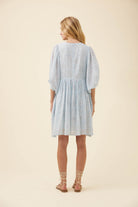 A woman viewed from the back wearing a light blue textured dress with puff sleeves and a gathered waist, paired with beige lace-up sandals, against a solid cream background.