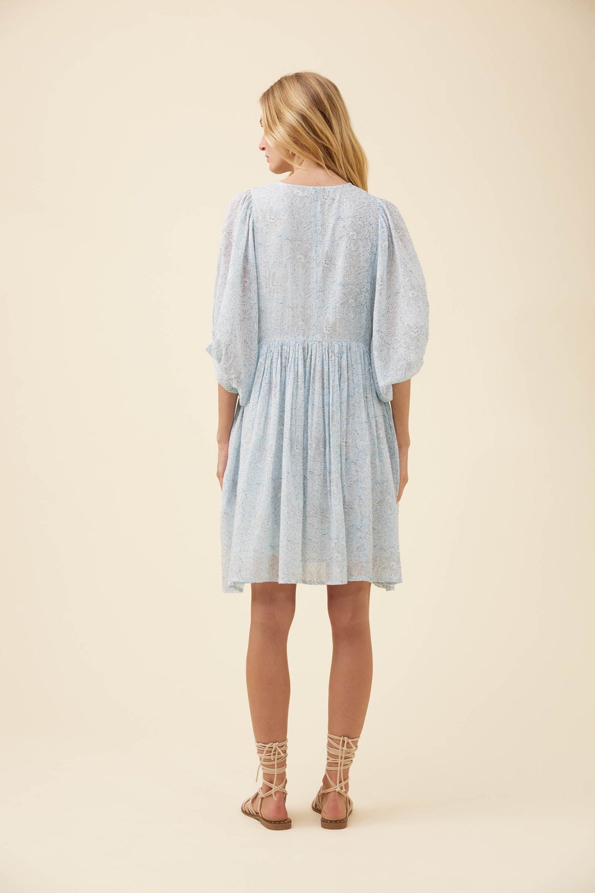 A woman viewed from the back wearing a light blue textured dress with puff sleeves and a gathered waist, paired with beige lace-up sandals, against a solid cream background.