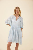 A woman modeling a light blue and white patterned short-sleeve dress with a gathered waist, standing against a solid beige studio background.