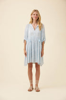 A woman modeling a light blue and white patterned short-sleeve dress with a V-neck and gathered waist, standing against a solid beige studio background.