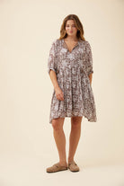 A woman modeling a short, loose-fitting dress with a brown and beige floral pattern, standing against a solid light beige studio background.