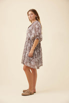 A woman modeling a short-sleeve, knee-length dress with a brown and beige botanical pattern, standing against a solid cream background.