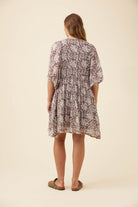 A woman viewed from behind wearing a short-sleeve, knee-length dress with a brown and beige floral pattern, paired with brown flat sandals, against a solid cream background.