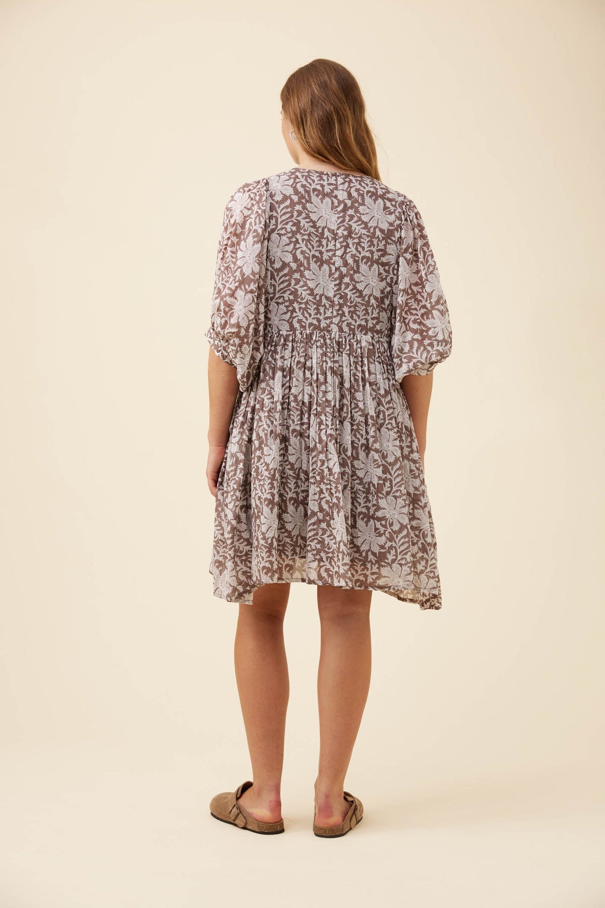 A woman viewed from behind wearing a short-sleeve, knee-length dress with a brown and beige floral pattern, paired with brown flat sandals, against a solid cream background.