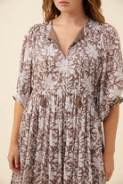 A woman wearing a brown and white floral print dress with a V-neckline, short puff sleeves, and tassel ties at the waist, shown against a plain beige background.