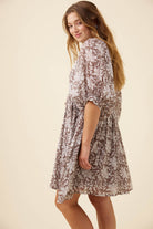 A woman wearing a brown and beige floral print dress, photographed from a side-back angle against a solid light beige studio background.