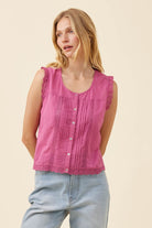 A woman wearing a sleeveless pink blouse with ruffle trim and a button-front bodice, paired with light blue jeans, against a plain beige studio background.