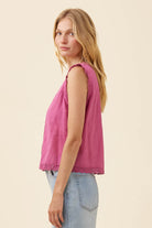 A woman shown in side profile wearing a sleeveless dusty pink top with delicate lace trim at the neckline and hem, paired with light blue jeans, against a solid cream background.