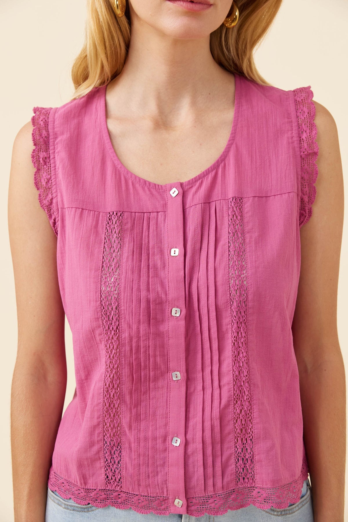 A sleeveless pink blouse with a scoop neckline, button-front center, vertical gathered pleats, and lace trim at the shoulders and hem, shown on a model against a light beige background.