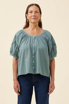 A woman modeling a dusty sage green short-sleeve blouse with puff sleeves, a gathered neckline, and a button-down front, paired with dark blue jeans against a solid cream background.