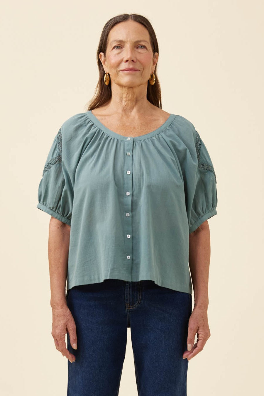 A woman modeling a dusty sage green short-sleeve blouse with puff sleeves, a gathered neckline, and a button-down front, paired with dark blue jeans against a solid cream background.