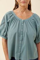A woman wearing a dusty sage green short-sleeve blouse with a gathered round neckline, front button closure, and lace detail on one sleeve, shown against a plain beige background.