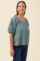A woman modeling a sage green short-sleeve blouse with puff sleeves, a gathered neckline, and button-front closure, paired with dark blue jeans against a plain beige studio background.