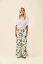 A woman stands against a plain beige studio background wearing a white puff-sleeve blouse and wide-leg trousers with a colorful paisley print.