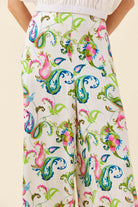 A close-up of a model wearing high-waisted, wide-leg trousers with a colorful paisley and floral print on an off-white base, paired with a white top.