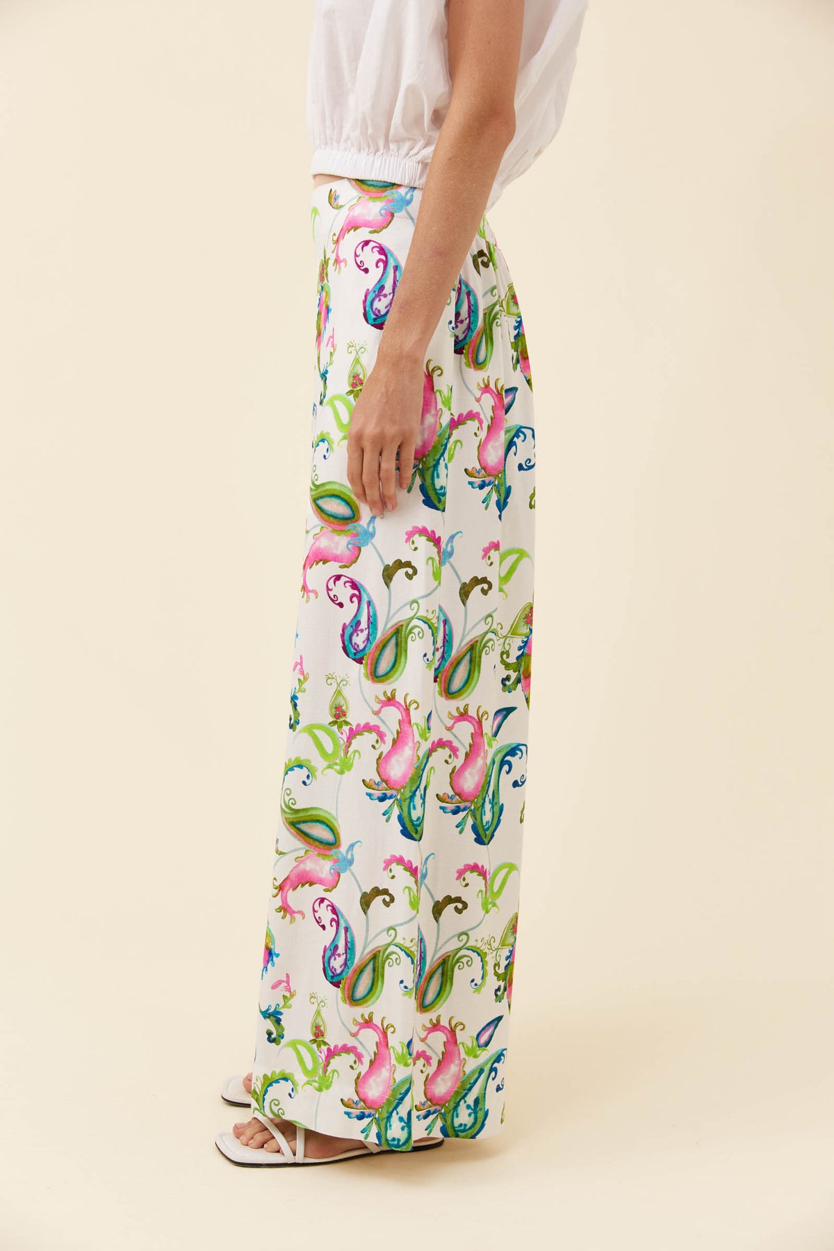 A model is shown from the waist down wearing white wide-leg trousers with a vibrant, colorful paisley pattern in pink, green, blue, and yellow, paired with a white top and sandals on a solid beige background.