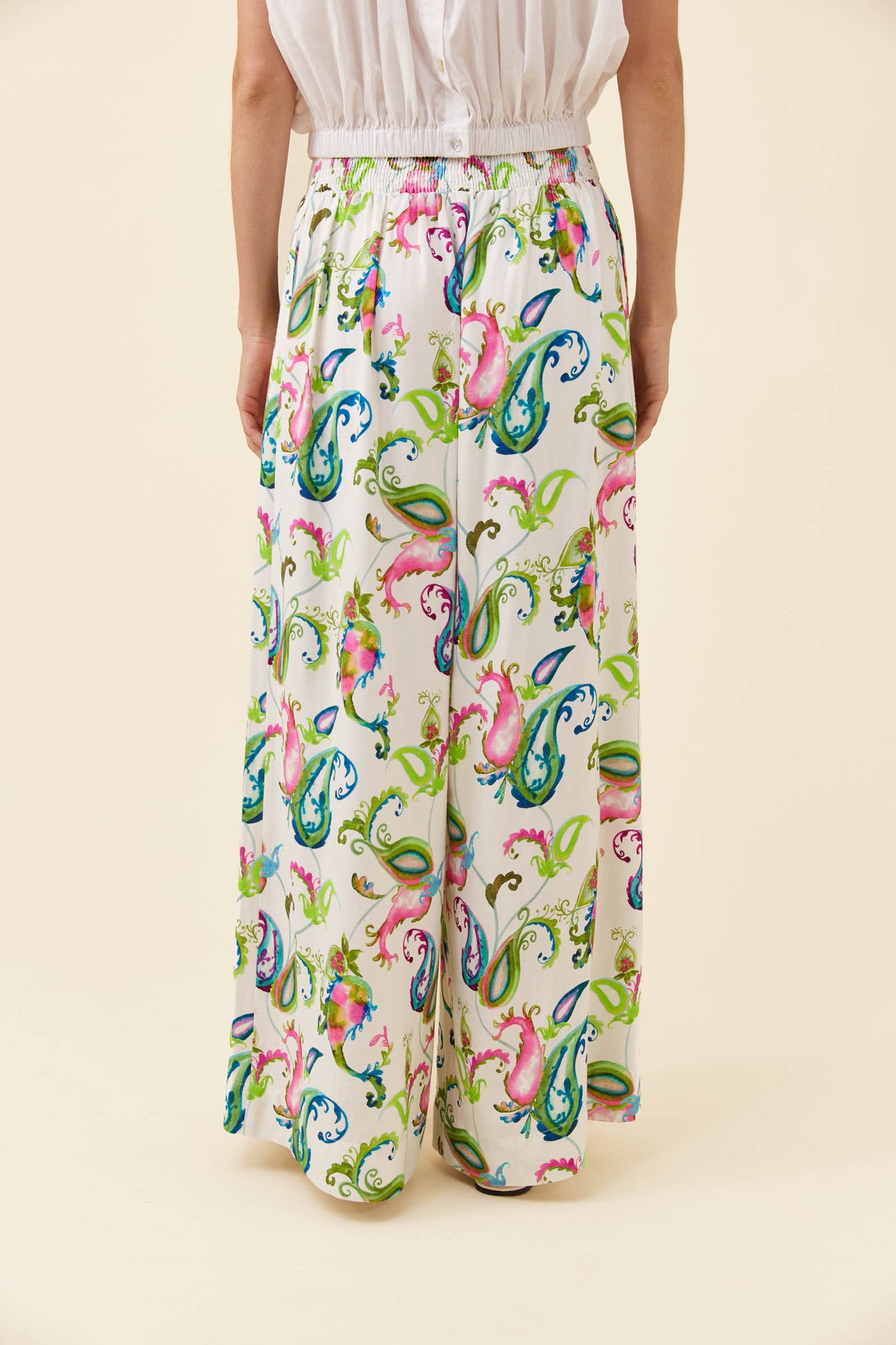 A model is shown from the waist down wearing high-waisted, wide-leg trousers with a vibrant multicolor paisley pattern on a cream background.