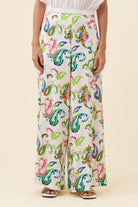 A person wearing high-waisted, wide-leg trousers with a colorful paisley and floral print on a cream background, paired with a white top and sandals, against a solid beige studio backdrop.
