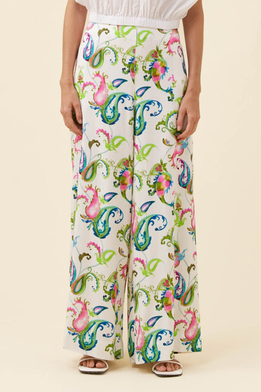 A person wearing high-waisted, wide-leg trousers with a colorful paisley and floral print on a cream background, paired with a white top and sandals, against a solid beige studio backdrop.