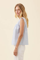 A woman wearing a light blue and white vertically striped sleeveless top with a gathered neckline, paired with white pants, photographed against a solid light beige background.