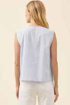 A woman shown from the back wearing a light blue and white vertically striped sleeveless top and white pants, set against a solid cream background.