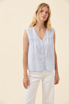 A woman modeling a sleeveless light blue and white vertically striped button-up blouse with a V-neck and gathered shoulders, paired with white pants, against a solid light beige background.
