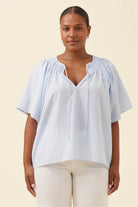A woman modeling a light blue and white vertically striped short-sleeve blouse with a smocked V-neckline, paired with cream pants, against a solid beige background.