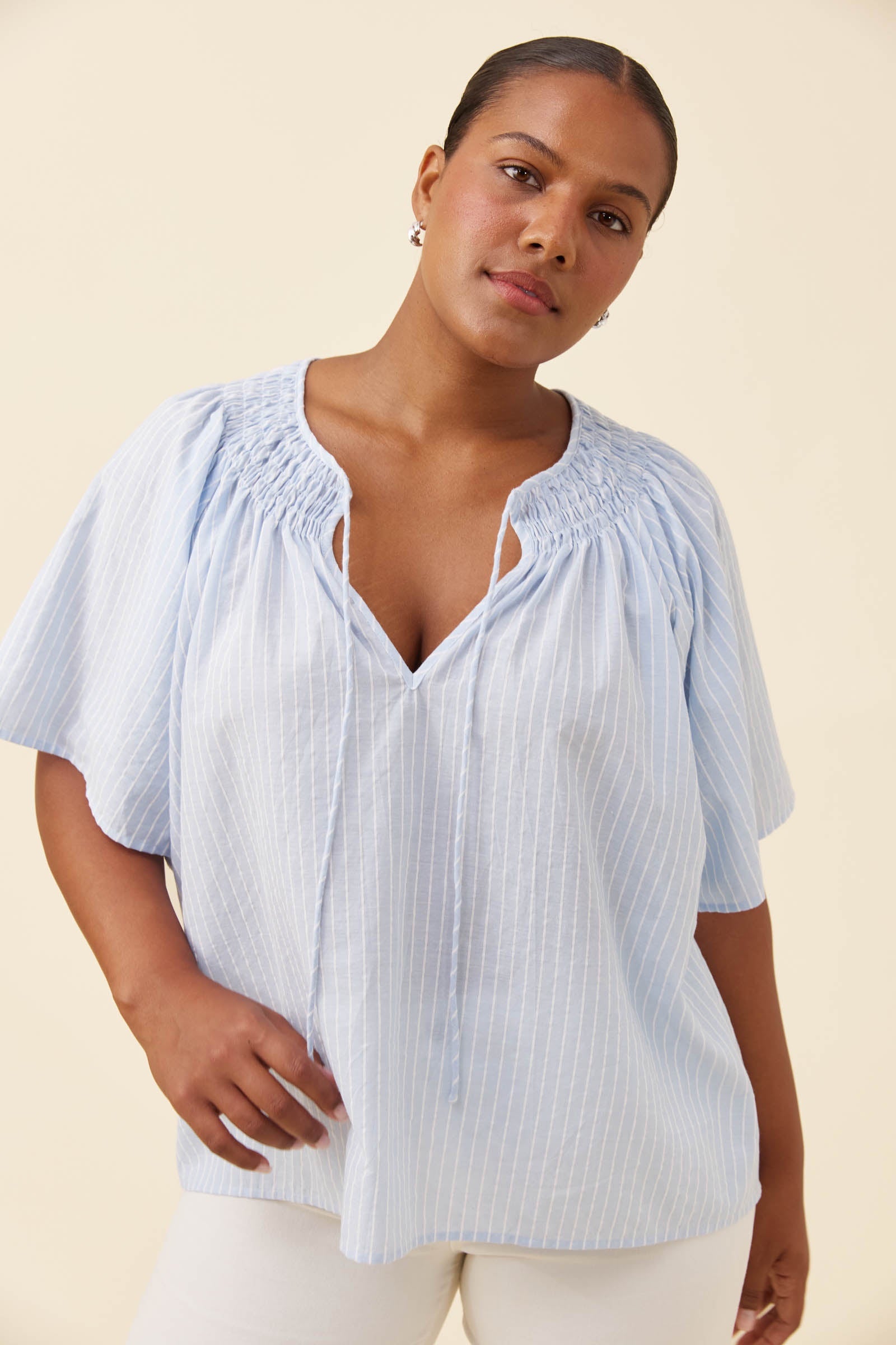 A woman wearing a light blue and white vertically striped short-sleeve blouse with a smocked neckline, paired with white pants, against a solid beige background.