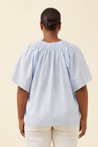 A woman viewed from the back wearing a light blue and white vertically striped smocked blouse with short puff sleeves, paired with cream pants, against a solid beige background.