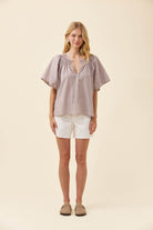 A woman stands against a plain beige studio background wearing a short-sleeve smocked blouse with vertical stripes in muted pink and beige, white shorts, and tan slip-on shoes.