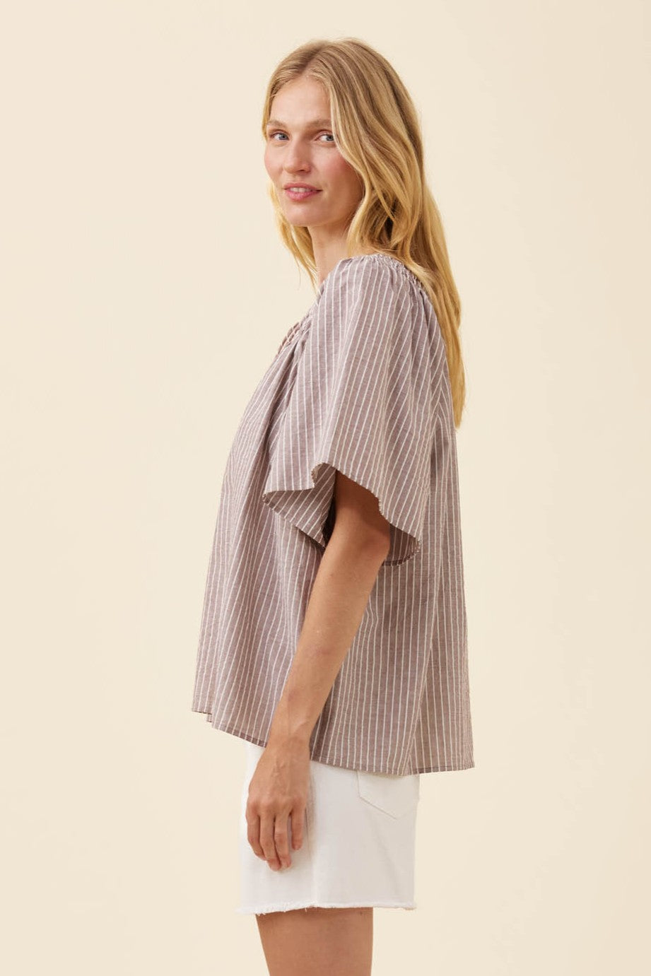 A woman modeling a loose, short-sleeve blouse with vertical stripes in beige and brown, paired with white shorts, against a solid light beige background.