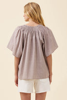 A woman shown from the back wearing a loose, short-sleeve smocked blouse with vertical stripes in beige and brown, paired with white shorts, against a solid light beige background.