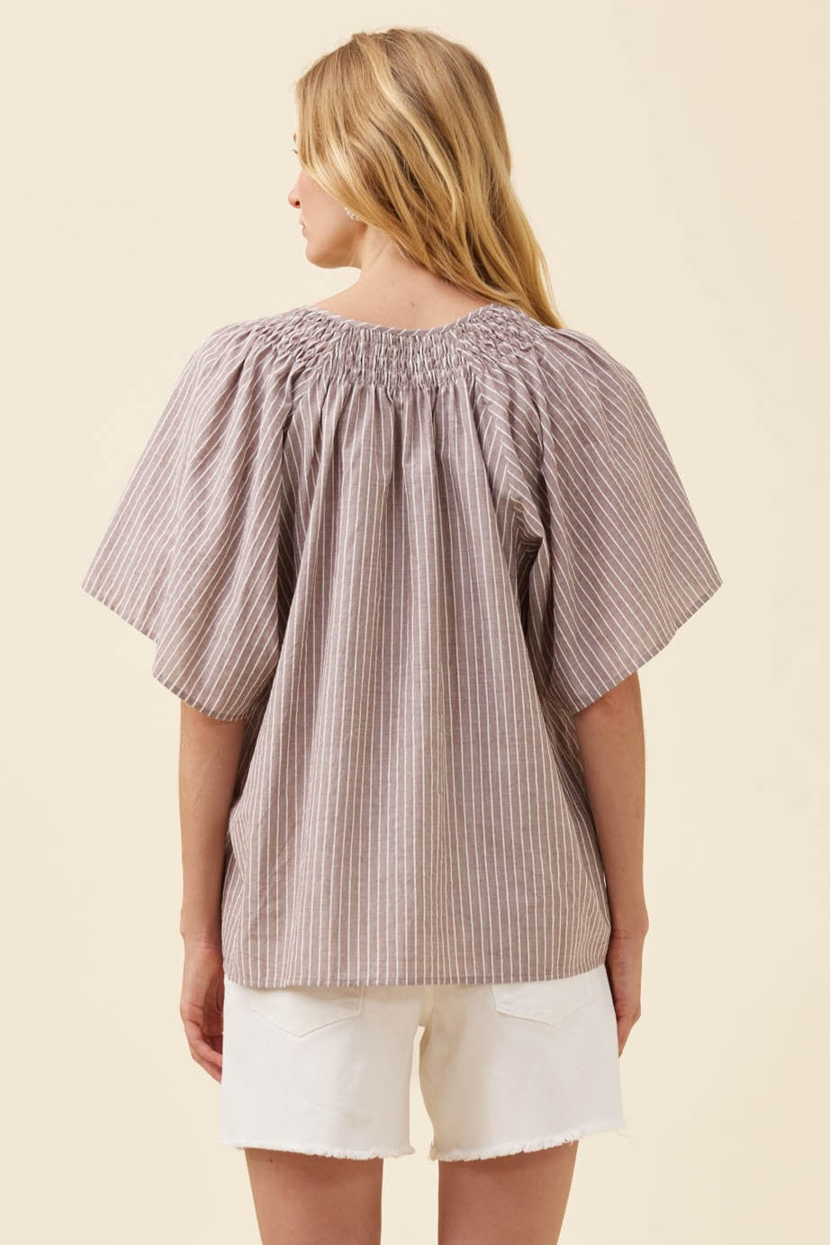 A woman shown from the back wearing a loose, short-sleeve smocked blouse with vertical stripes in beige and brown, paired with white shorts, against a solid light beige background.