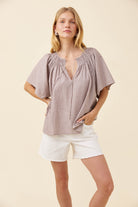 A woman modeling a short-sleeve smocked blouse with vertical stripes in a muted taupe-brown and cream color, paired with white shorts, against a solid beige background.