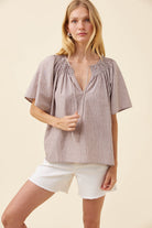A woman modeling a short-sleeve smocked blouse with vertical stripes in a muted taupe-brown and beige color, paired with white shorts, against a solid cream background.