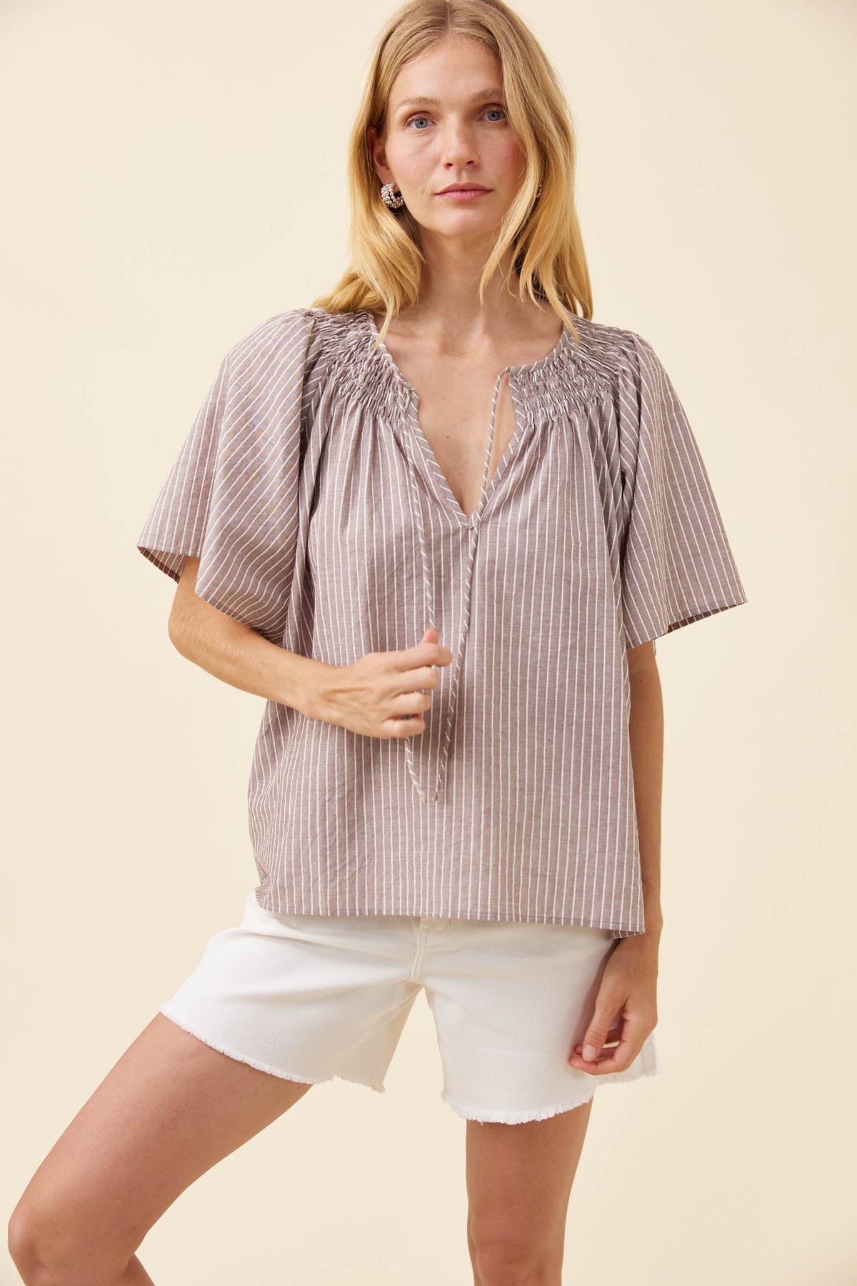 A woman modeling a short-sleeve smocked blouse with vertical stripes in a muted taupe-brown and beige color, paired with white shorts, against a solid cream background.