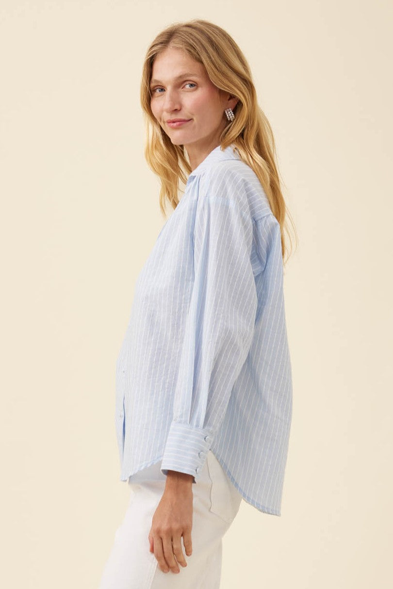 A woman wearing a light blue and white vertically striped button-down shirt with white pants, photographed against a solid light beige studio background.