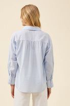 A woman shown from the back wearing a light blue and white vertically striped button-up shirt with a gathered back seam, paired with white pants, against a solid beige background.
