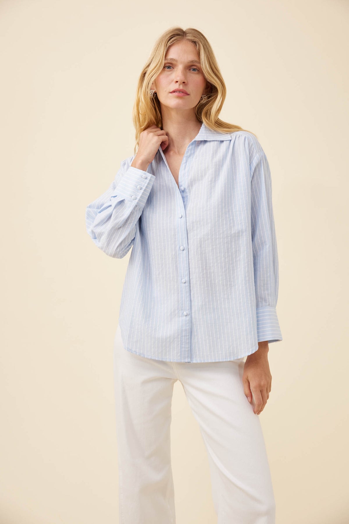 A woman modeling a light blue and white vertically striped button-down shirt with long sleeves, paired with white pants, against a solid beige studio background.