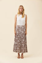 A woman stands against a plain beige studio background wearing a sleeveless white top and a long pleated skirt with a brown and beige floral pattern.
