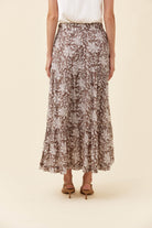 A woman wearing a brown and beige floral print maxi skirt with a white top and brown heeled sandals, shown from the waist down against a solid cream background.