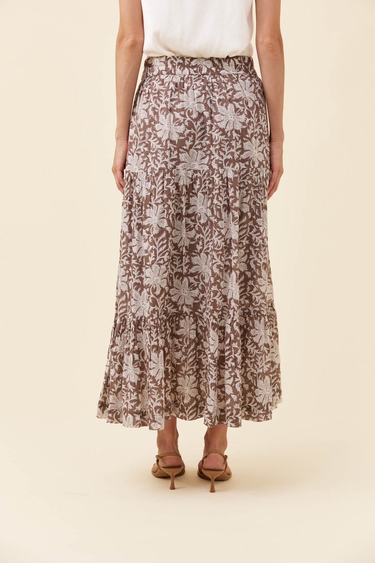 A woman wearing a brown and beige floral print maxi skirt with a white top and brown heeled sandals, shown from the waist down against a solid cream background.