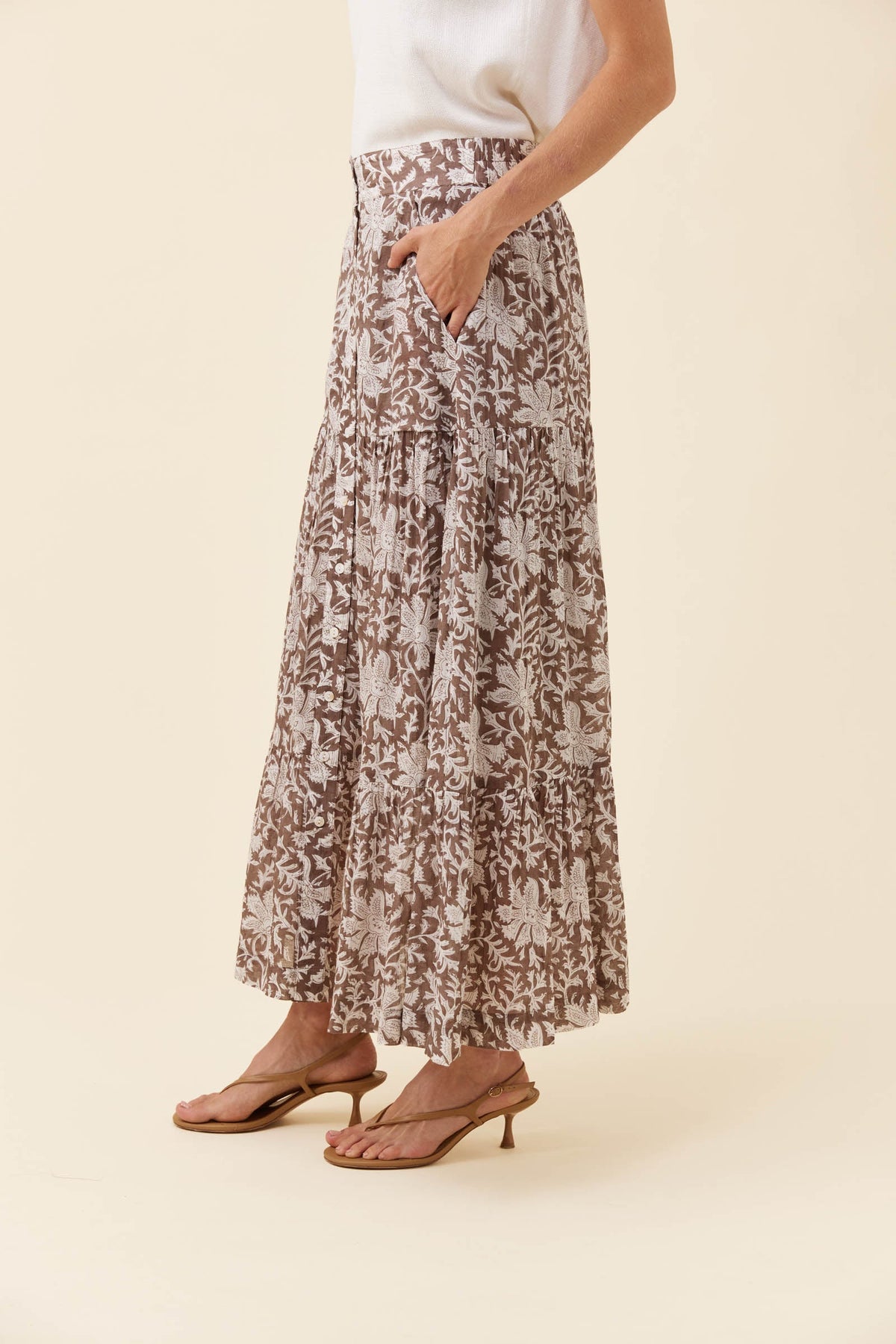 A model wearing a brown and beige floral print maxi skirt with pockets, paired with a white top and brown heeled sandals, against a solid cream background.