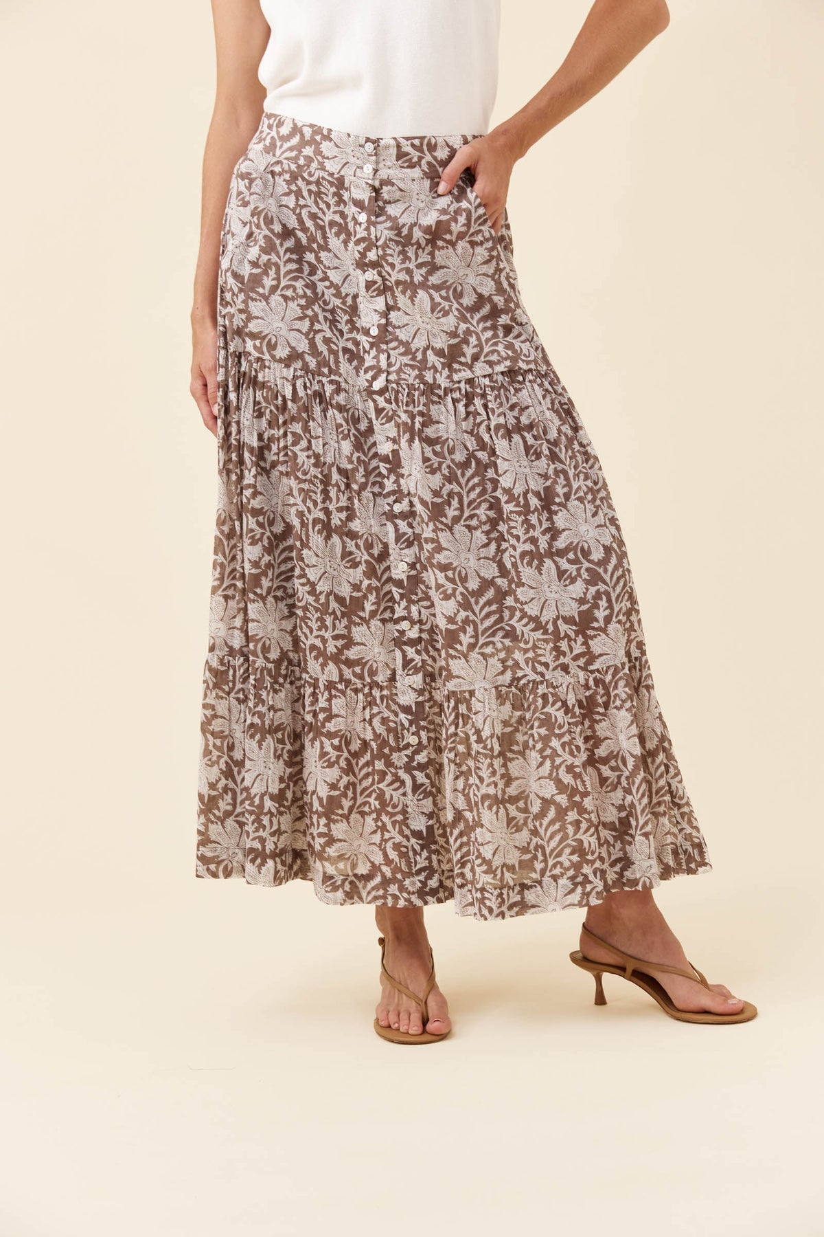 A woman wearing a long, tiered maxi skirt with a brown and beige floral pattern, paired with a white top and brown heeled sandals, against a solid beige background.