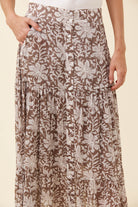 A close-up of a person wearing a brown and beige floral print skirt with a button-front waistband and pleated tiers, against a plain light beige background.